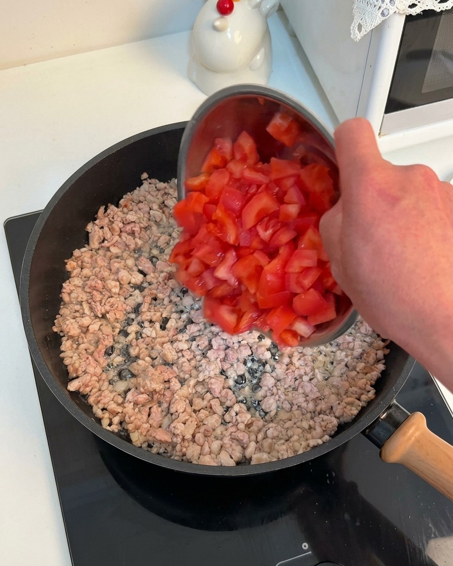 Una mano che versa una ciotola di pomodori rossi a cubetti in una padella con carne macinata rosolata.