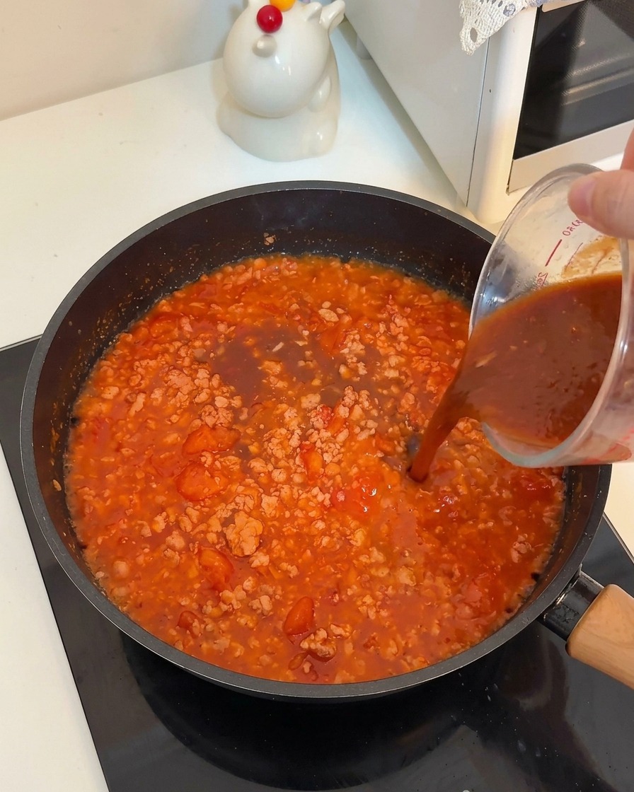 Una salsa liquida scura versata da un misurino di vetro in una padella con carne e pomodori che sobbollono.
