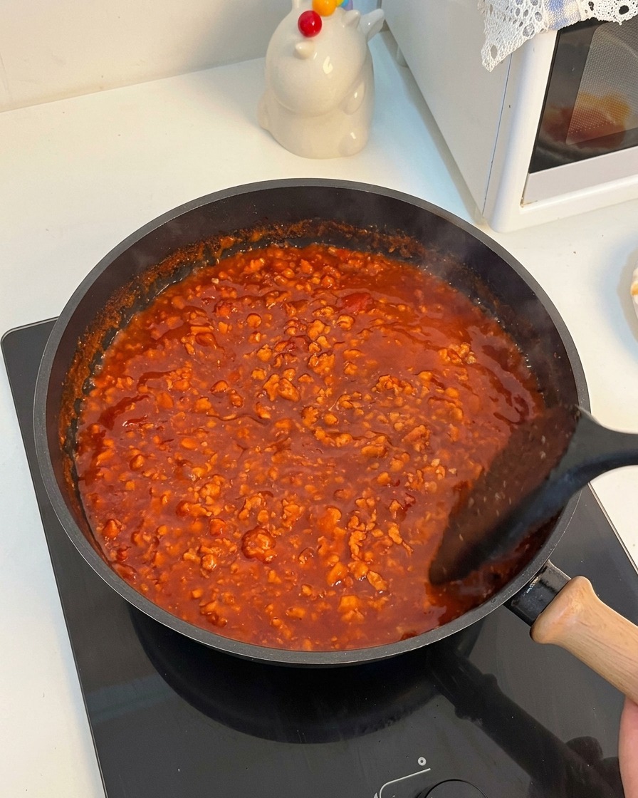 Ragù di carne e pomodoro vibrante che bolle vivacemente in una padella mentre viene mescolato con una spatola nera.