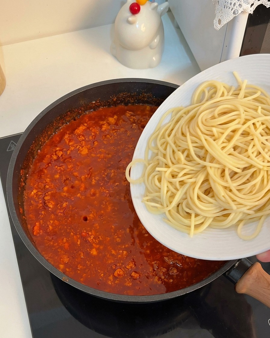 Spaghetti cotti che vengono fatti scivolare da un piatto bianco in una padella piena di ricco ragù di carne.