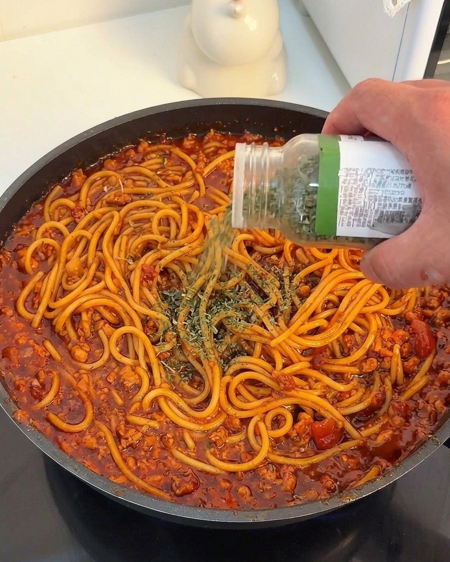 Una mano con un barattolo di spezie che spolvera prezzemolo secco su spaghetti conditi con ragù di carne rosso.