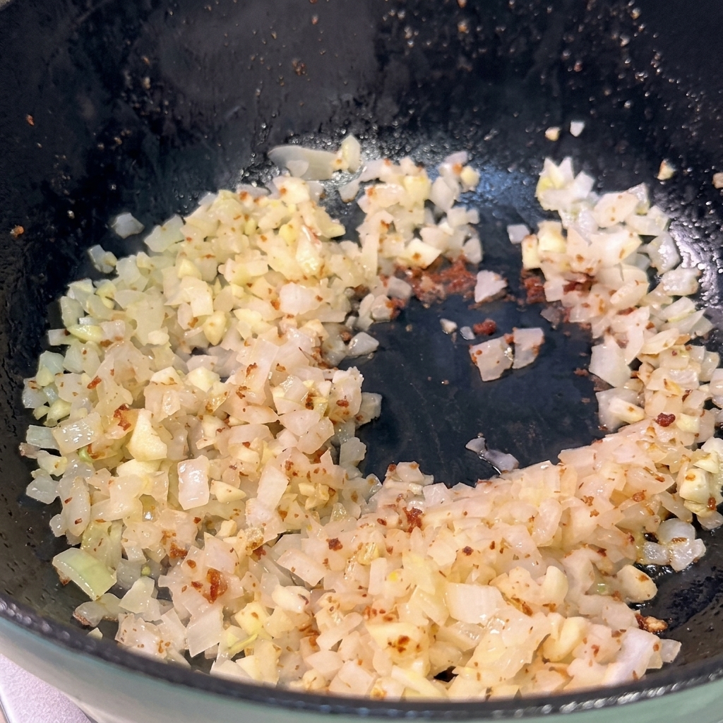 Cipolla e aglio tritati che soffriggono in una padella scura, diventando traslucidi.