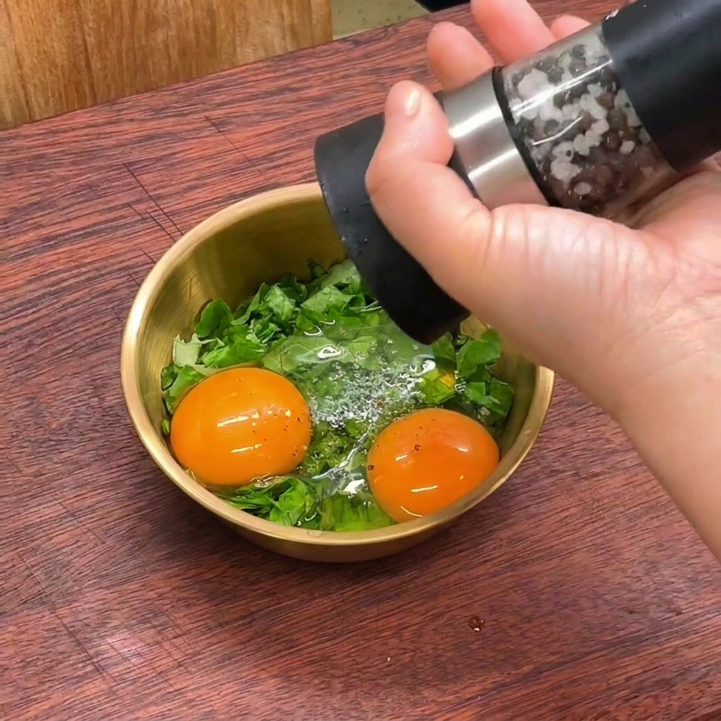 Una mano usa un macinapepe per macinare pepe nero fresco in una ciotola dorata contenente uova crude e lattuga tagliata.