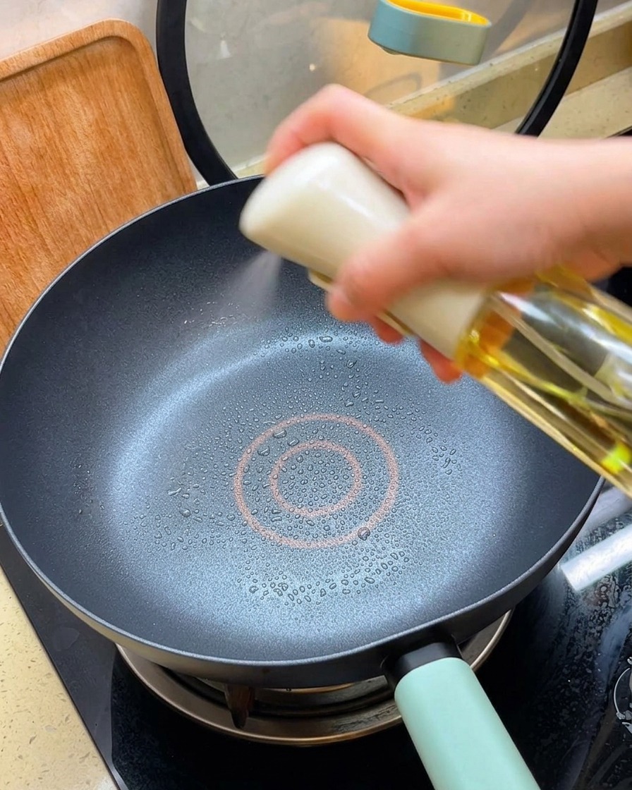 Una mano tiene uno spray per olio, nebulizzando un sottile strato di olio in una padella antiaderente nera riscaldata.