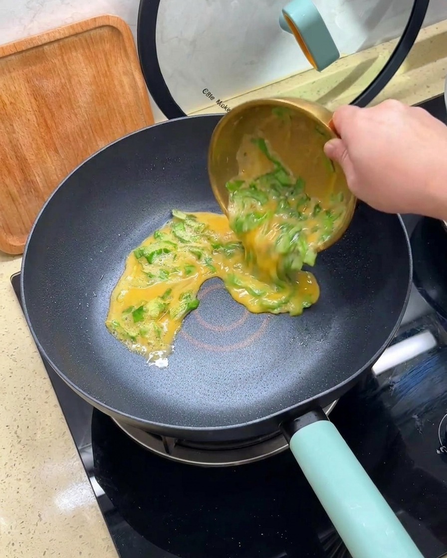 Versamento di un composto di uova sbattute e lattuga tritata da una ciotola dorata in una padella antiaderente calda.