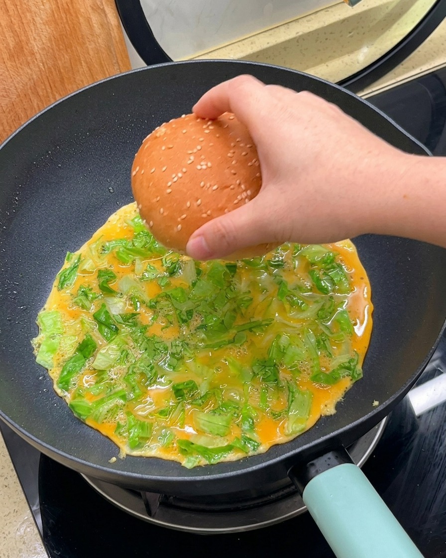 Una mano appoggia un pane da burger al sesamo rivolto verso il basso sopra il composto di uova e lattuga in cottura.