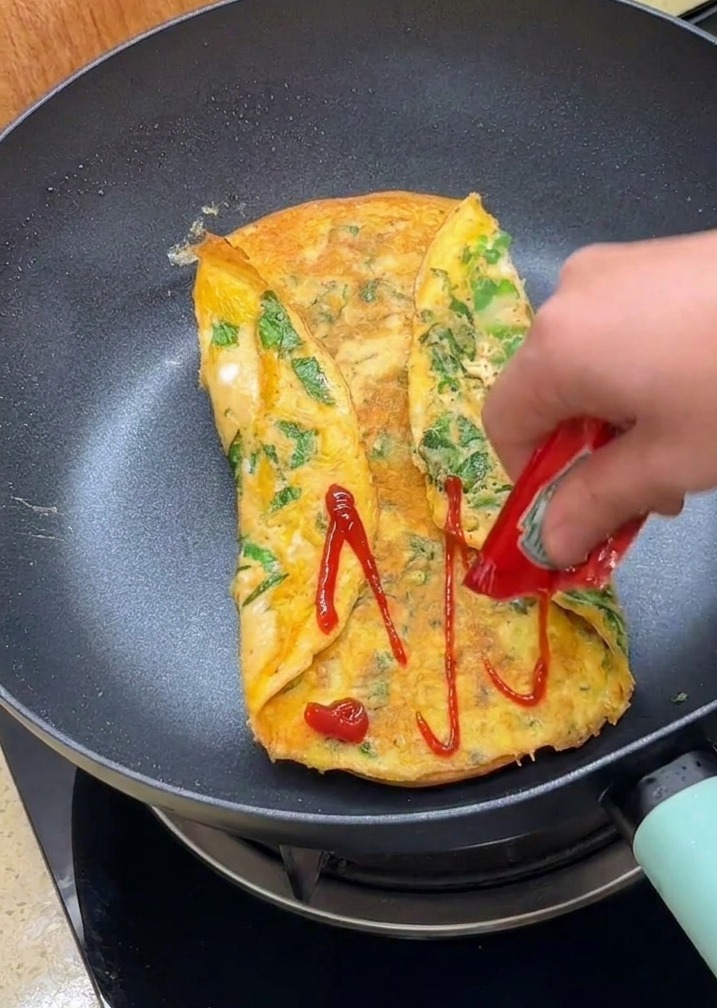 Una mano che spreme ketchup rosso da una bustina sulla superficie delluovo ripiegato in padella.