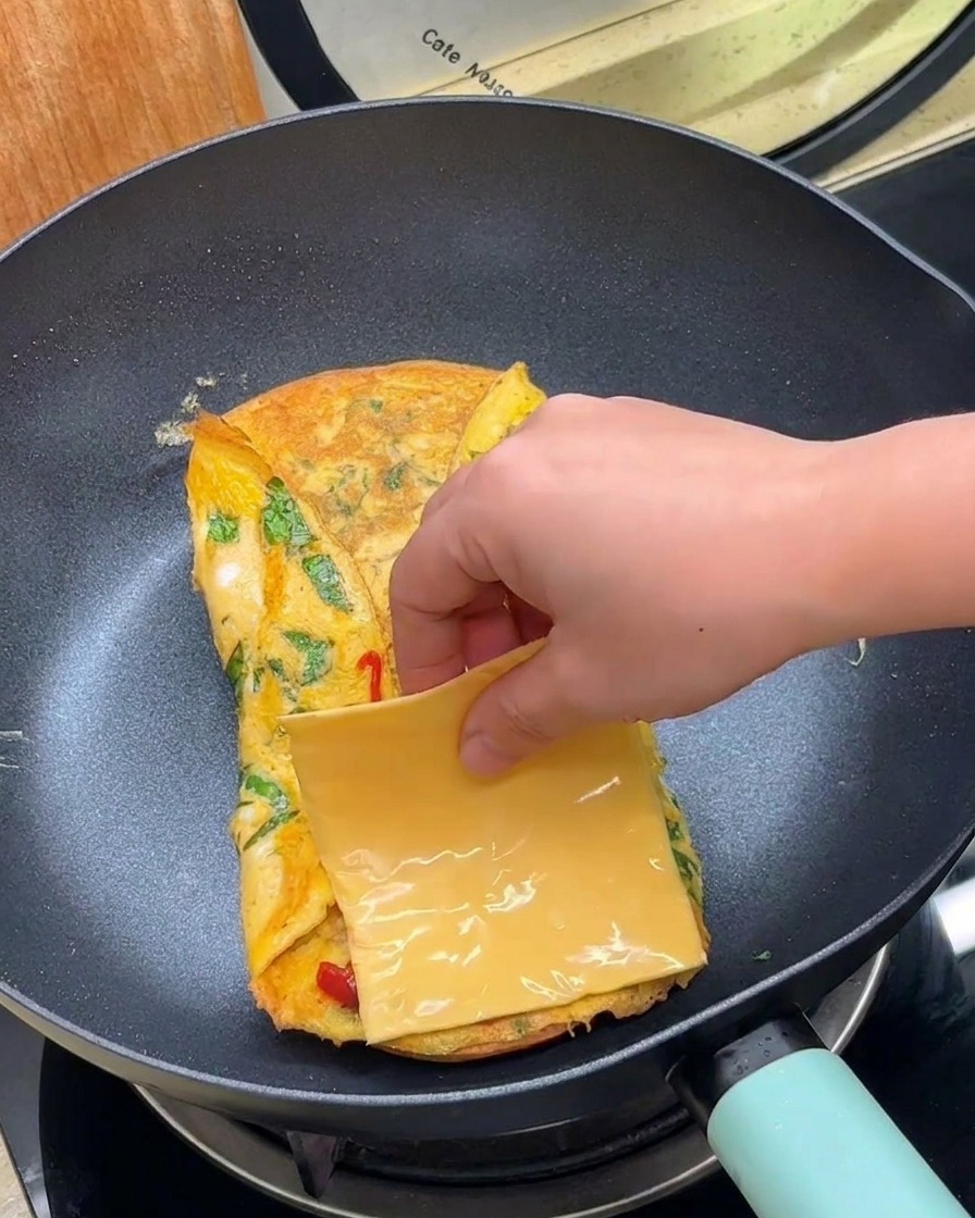 Una mano che posiziona una fetta quadrata di formaggio giallo su un uovo ripiegato in padella.