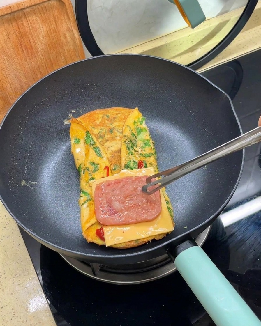 Pinze che posizionano una fetta di carne cotta sopra una fetta di formaggio su unomelette in padella.