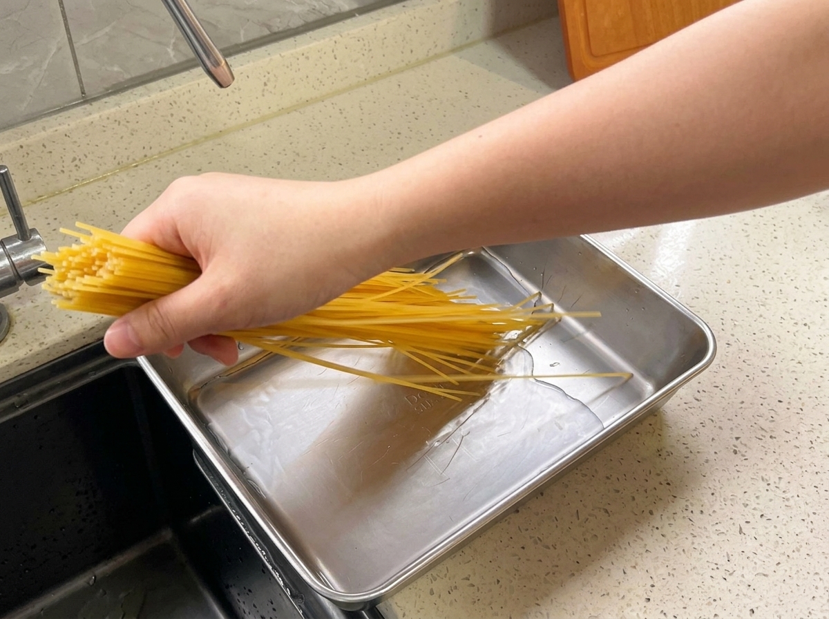 Una mano che immerge un fascio di spaghetti secchi in una teglia rettangolare di metallo piena dacqua.