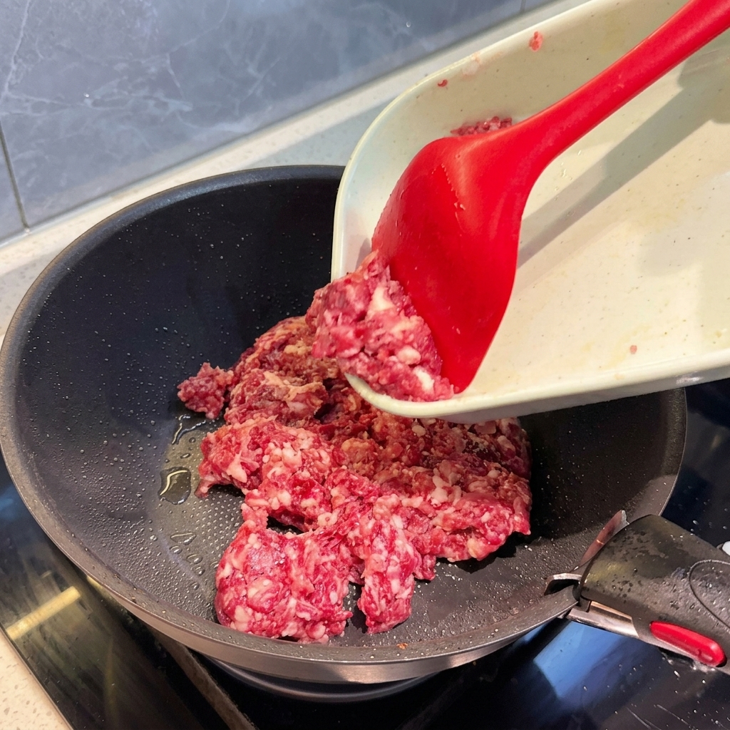 Carne macinata mista versata da un tagliere in una padella scura usando una spatola rossa.