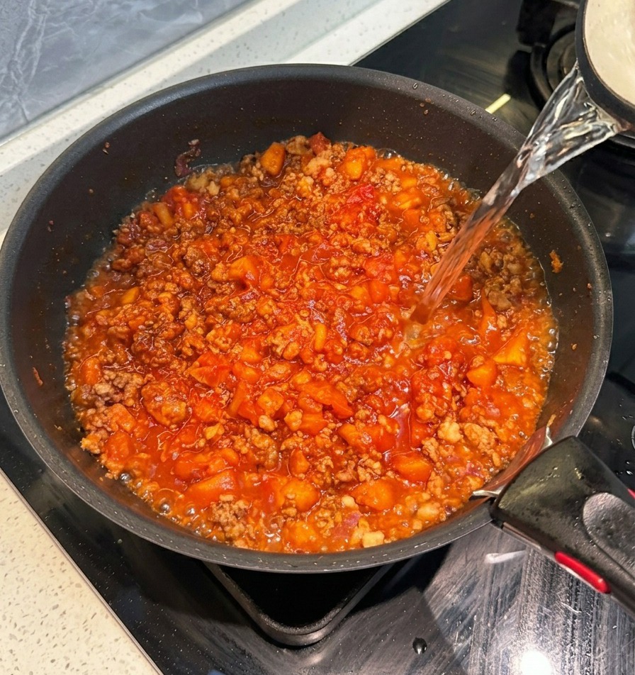 Acqua calda versata da un bollitore in una padella nera contenente un denso sugo rosso di carne.