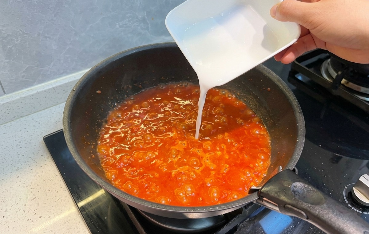 Una pastella di amido bianca versata da una ciotolina in un sugo di pomodoro bollente.