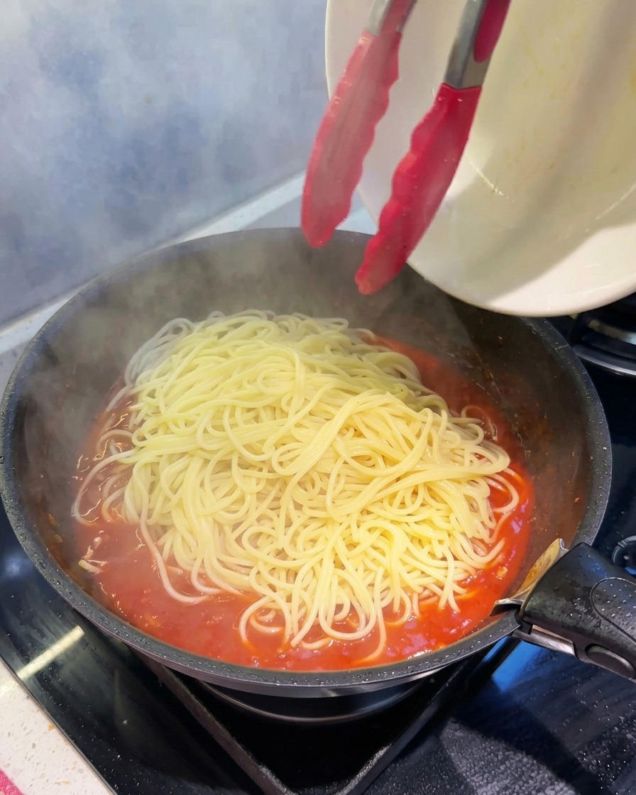 Pinze da cucina che trasferiscono spaghetti fumanti in una padella piena di denso ragù rosso.