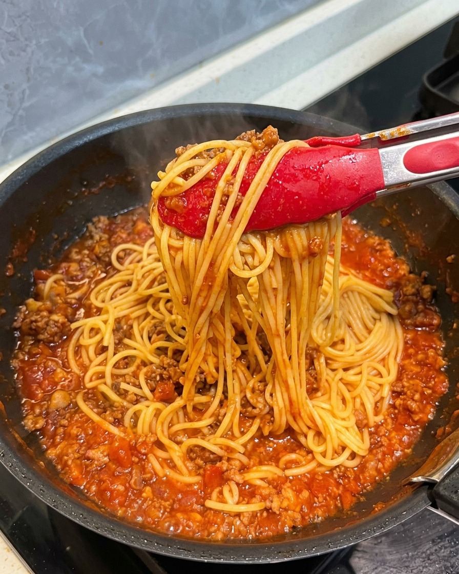 Pinze rosse che sollevano un nido di spaghetti completamente ricoperti da un ricco ragù di carne.