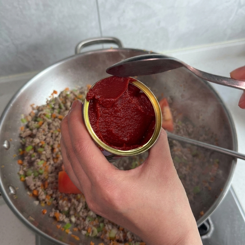 Una persona tiene un barattolo aperto di concentrato di pomodoro, usando un cucchiaio per versarlo nel wok.