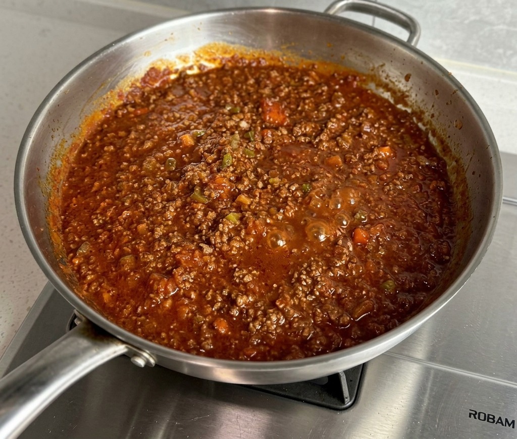 Ragù di carne rosso e denso che sobbolle in una padella di acciaio inox.