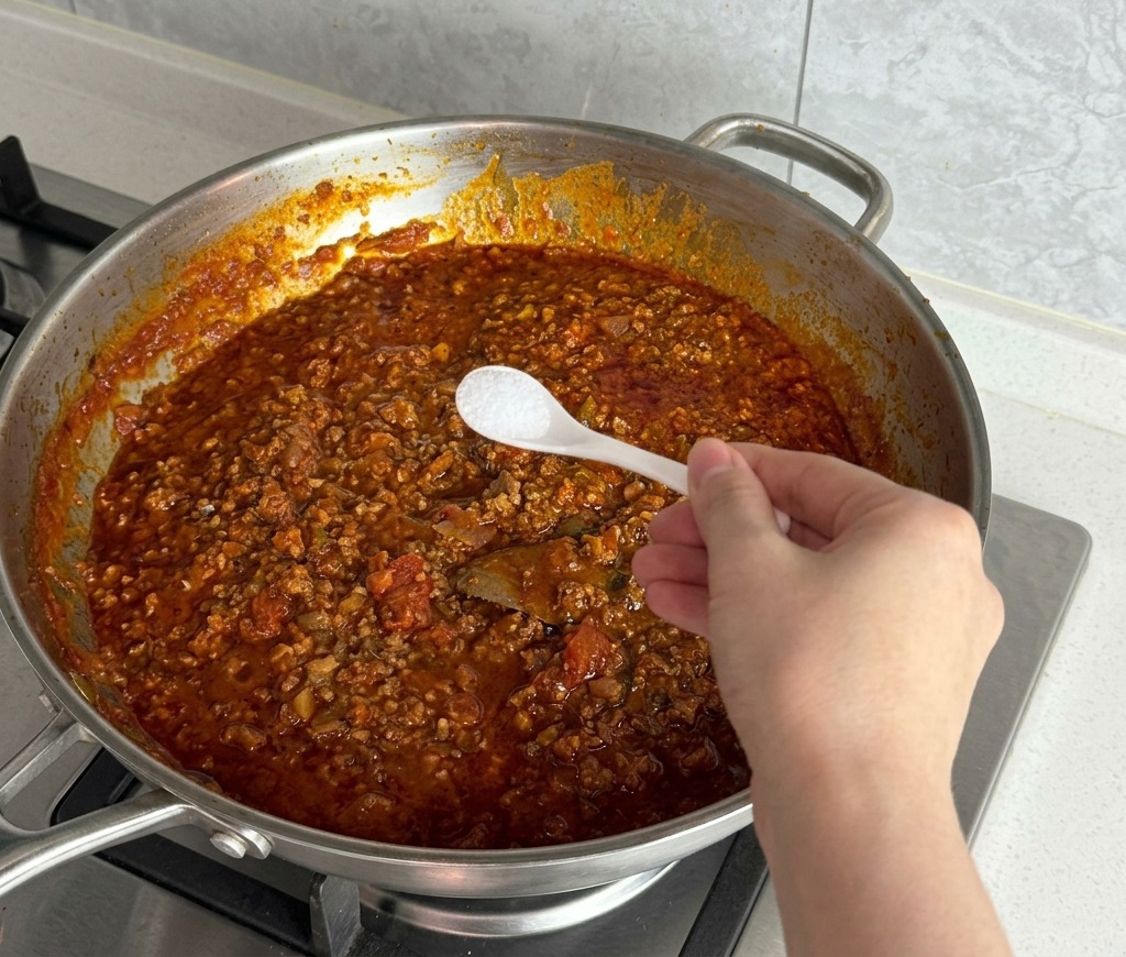 Una mano con un cucchiaino aggiunge sale bianco al ragù fumante.