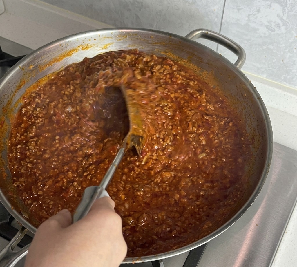 Una mano usa una spatola di metallo per mescolare energicamente il ragù denso in padella.