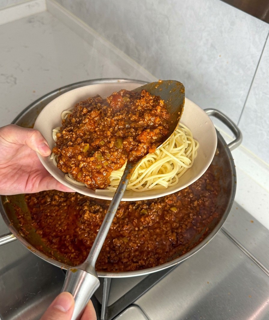 Un mestolo di ragù rosso e denso servito sopra una ciotola di spaghetti appena cotti.