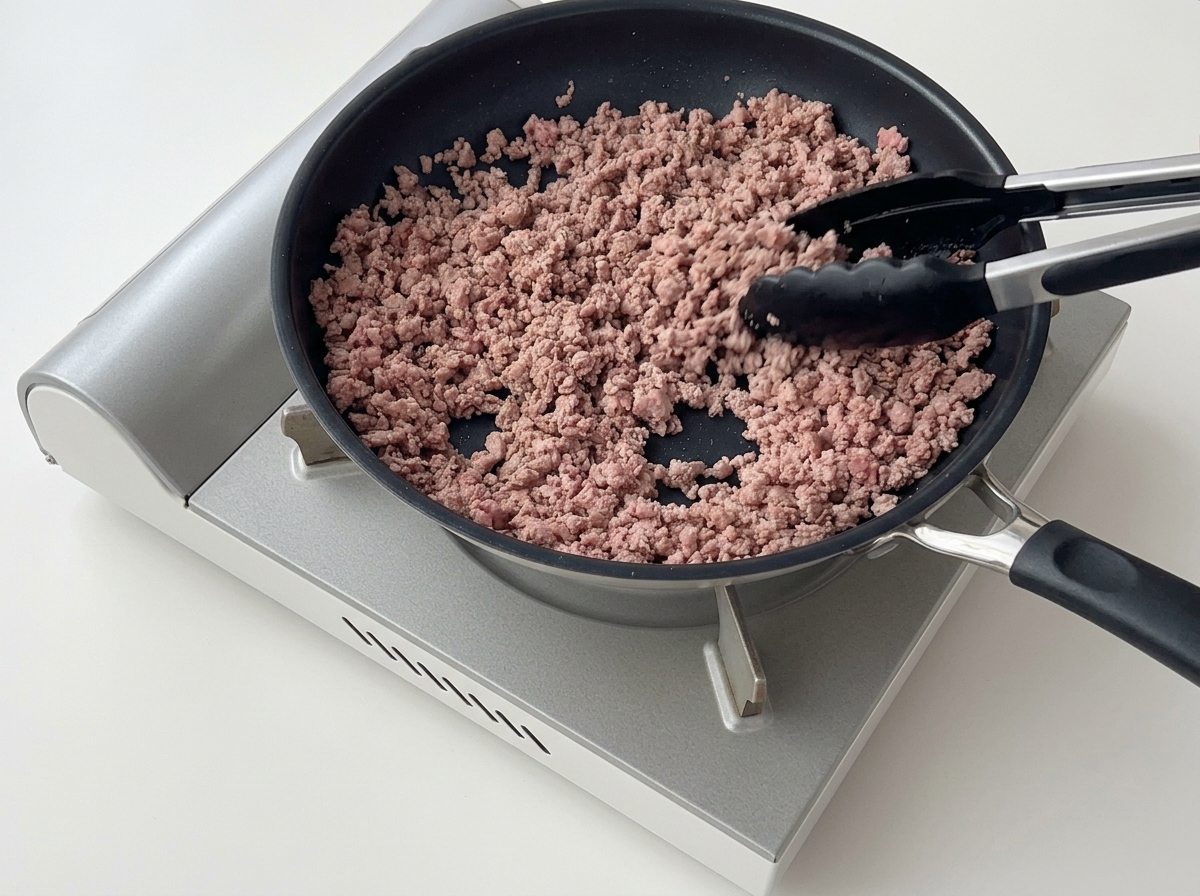 Pinze che sgranano e mescolano carne macinata rosolata in una padella antiaderente.