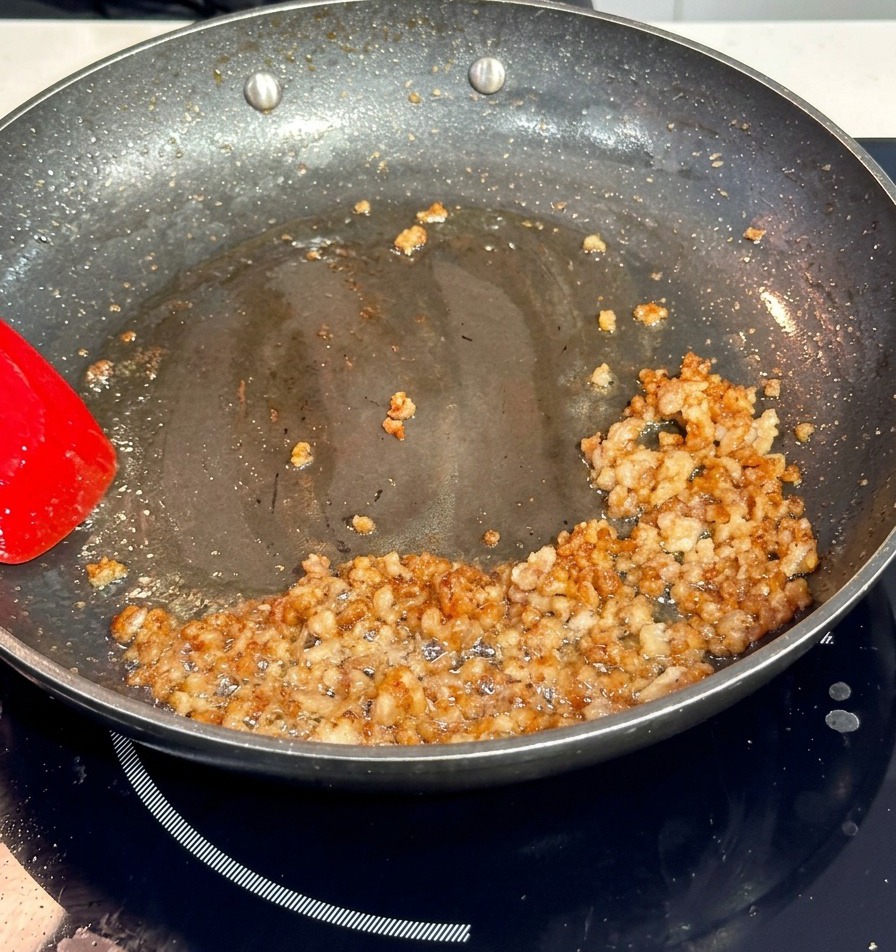 Carne di maiale che rosola in una padella antiaderente scura, mescolata con una spatola rossa.