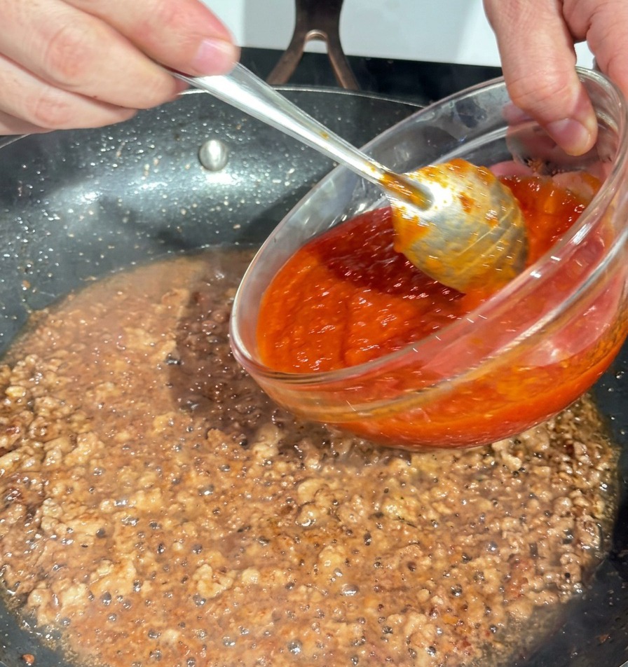 Sugo di pomodoro denso versato da una ciotola di vetro nella padella con il macinato.