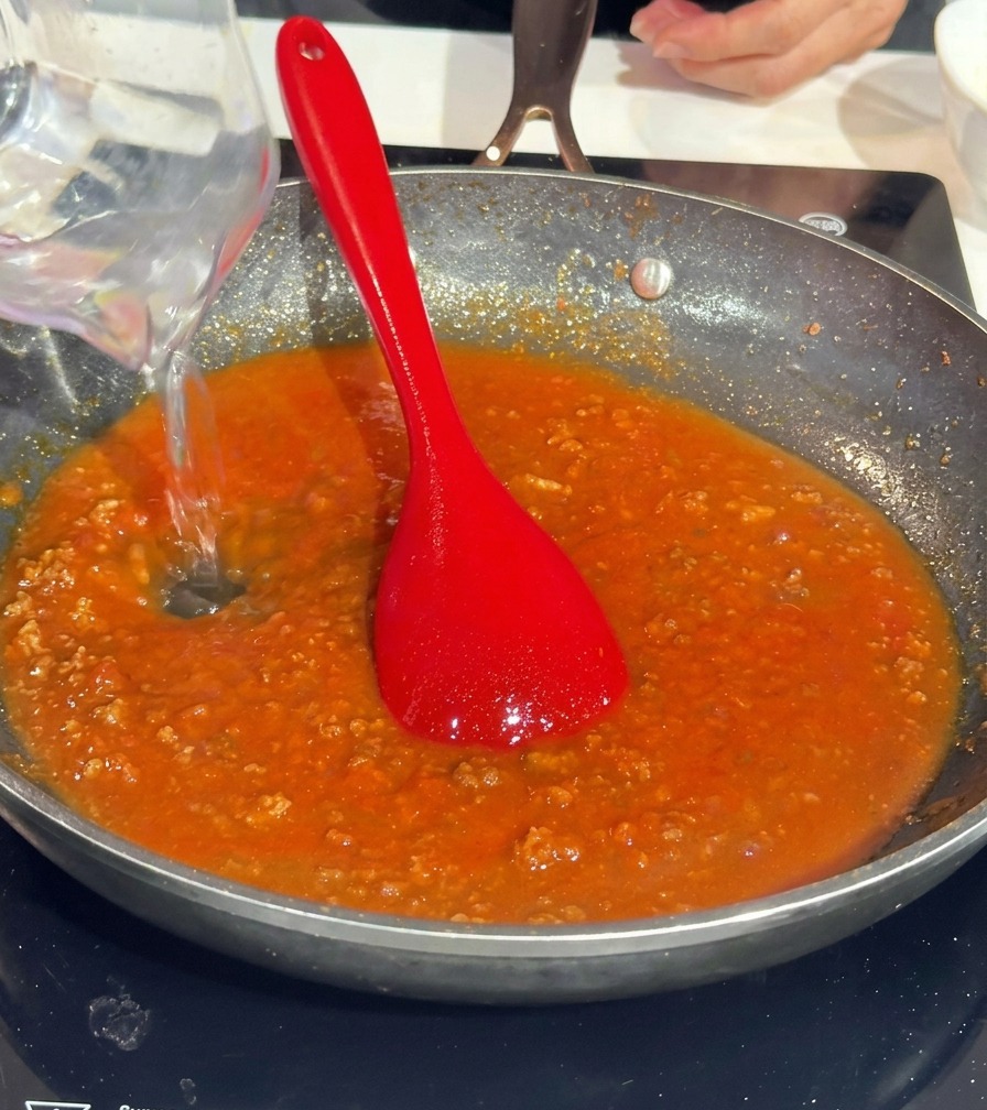 Acqua versata da un misurino trasparente in una padella con sugo rosso di carne.