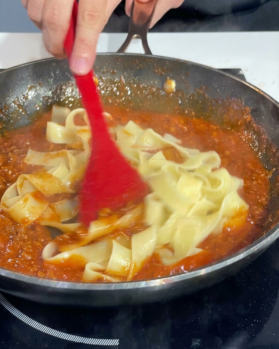 Spatola rossa che salta le pappardelle in padella per condirle uniformemente.
