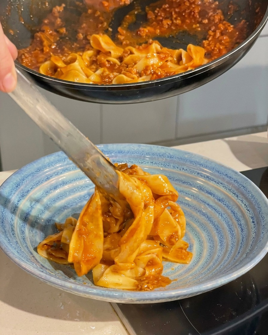 Pinze da cucina che sollevano la pasta condita dalla padella per adagiarla in una ciotola blu.