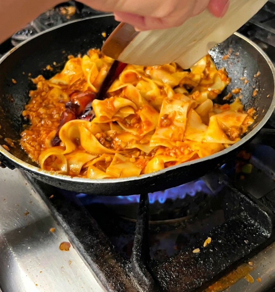 Salsa scura versata da un piattino sopra le pappardelle in padella.