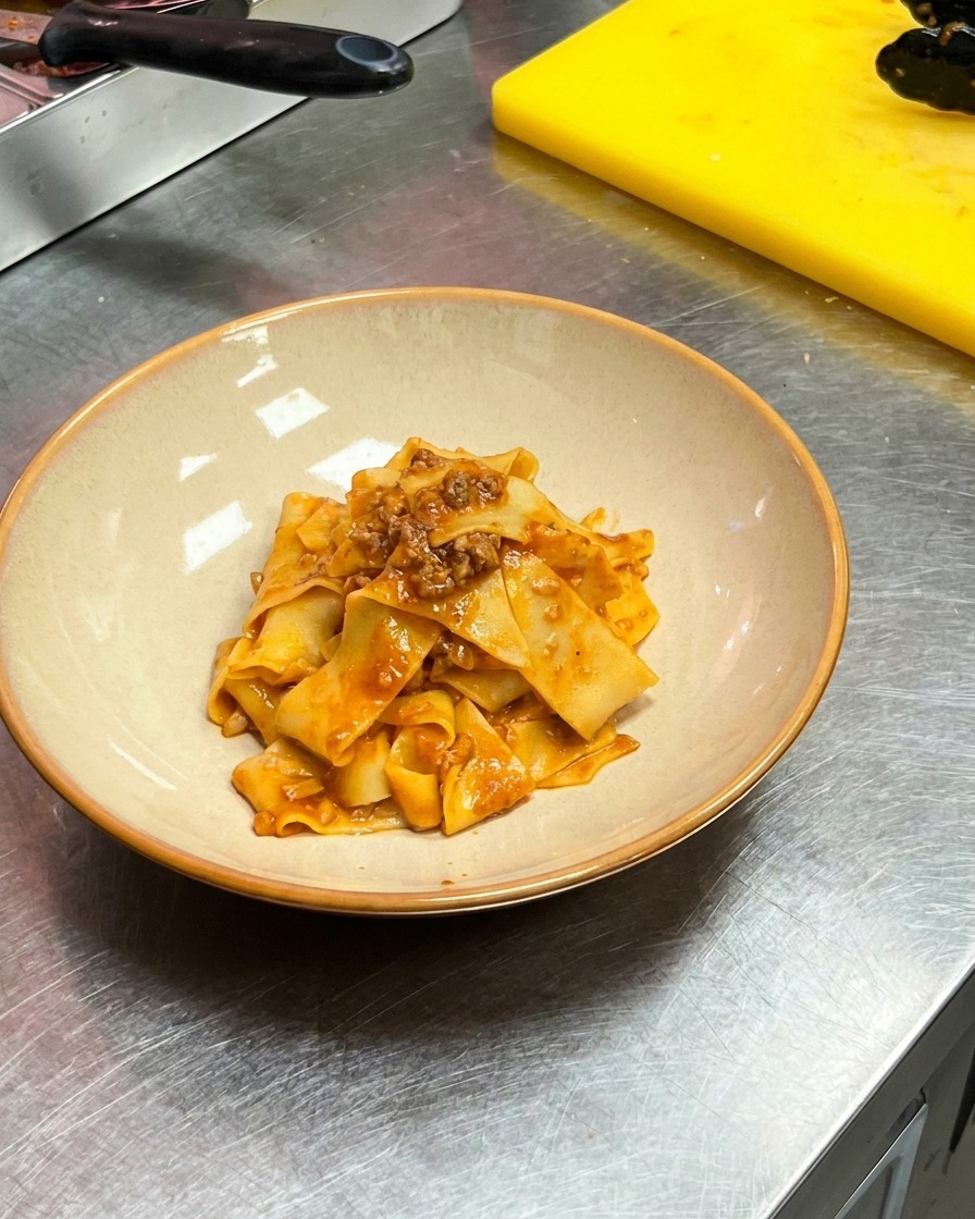 Pinze che trasferiscono le pappardelle al ragù in una ciotola di ceramica bianca.