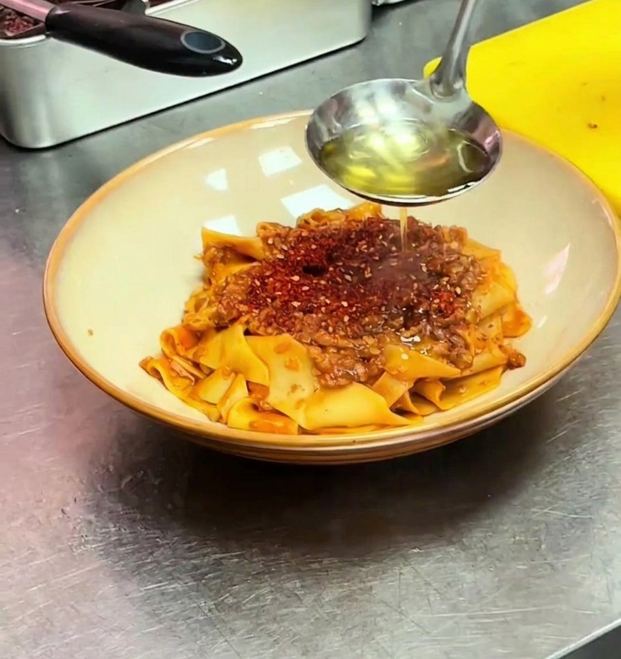 Un mestolo versa olio bollente sul peperoncino rosso sopra le pappardelle nella ciotola.