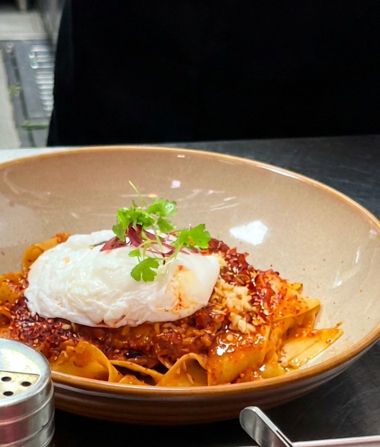 Ciotola finita di pappardelle piccanti con uovo in camicia, olio e micro-germogli.