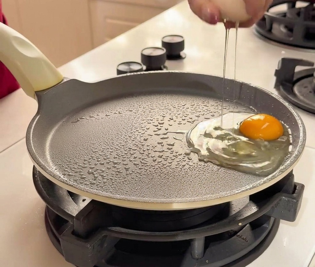 Una mano che rompe un uovo fresco in una padella antiaderente accanto a un altro uovo già aperto.