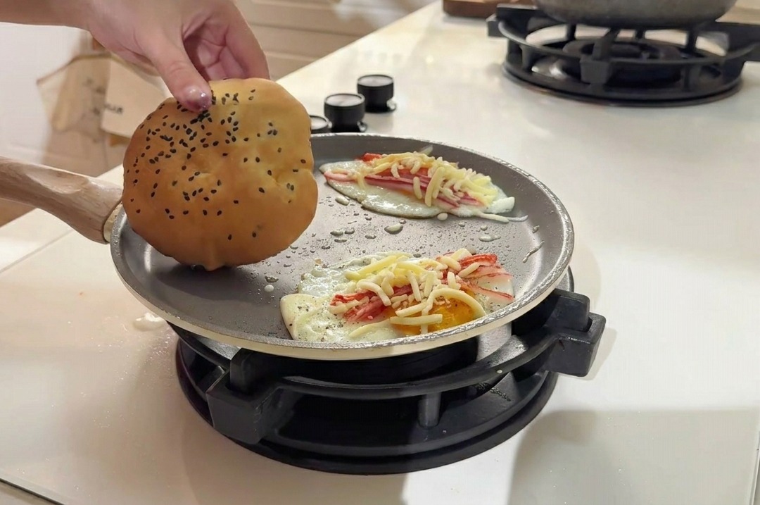 Un panino da burger dorato con semi di sesamo appoggiato sulla superficie della padella accanto alle uova.
