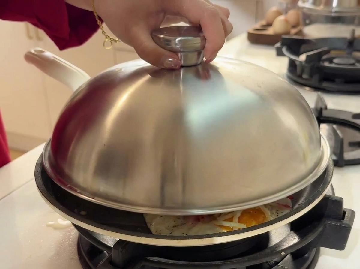 Una mano che posiziona un coperchio a cupola in acciaio inox sopra la padella con uova, formaggio e granchio.