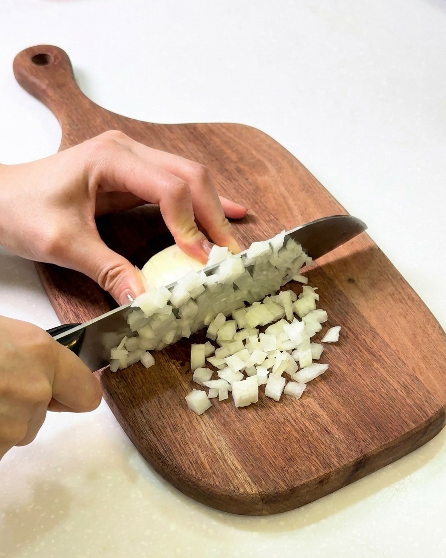 Mani che tritano finemente una cipolla bianca su un tagliere di legno.