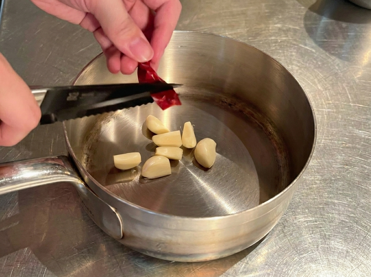Mani che tagliano un peperoncino rosso secco sopra il pentolino con gli spicchi daglio.