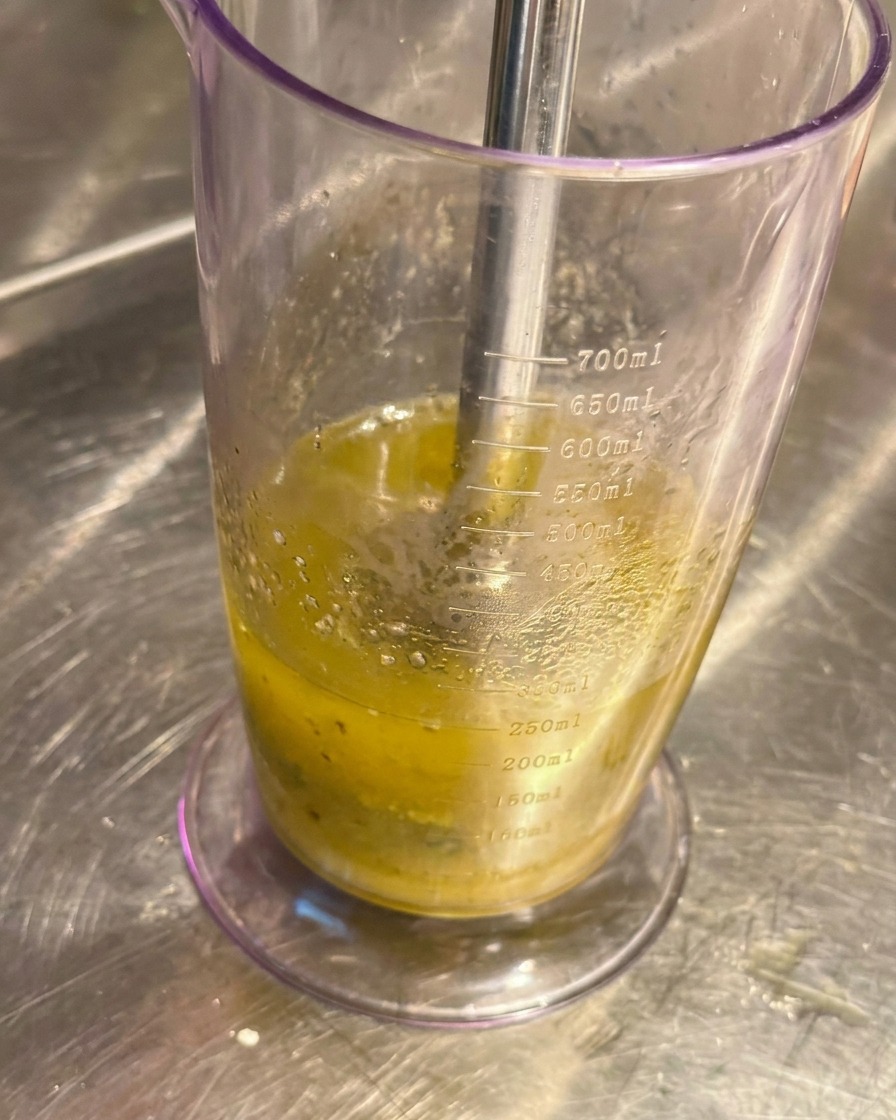 Frullatore a immersione in un bicchiere graduato trasparente che trasforma aglio e olio in una crema chiara.