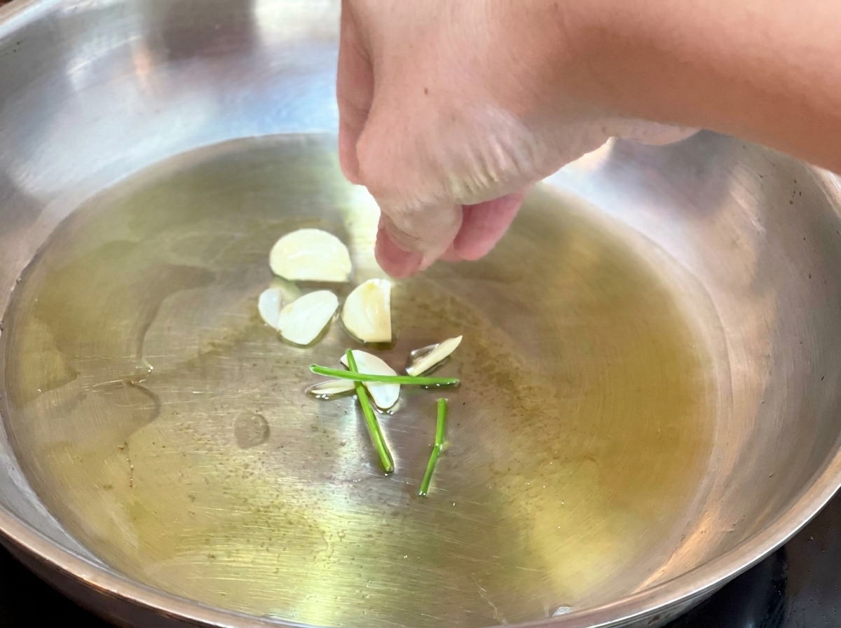 Mano che aggiunge aglio a fette e gambi di prezzemolo verde nellolio caldo in padella.