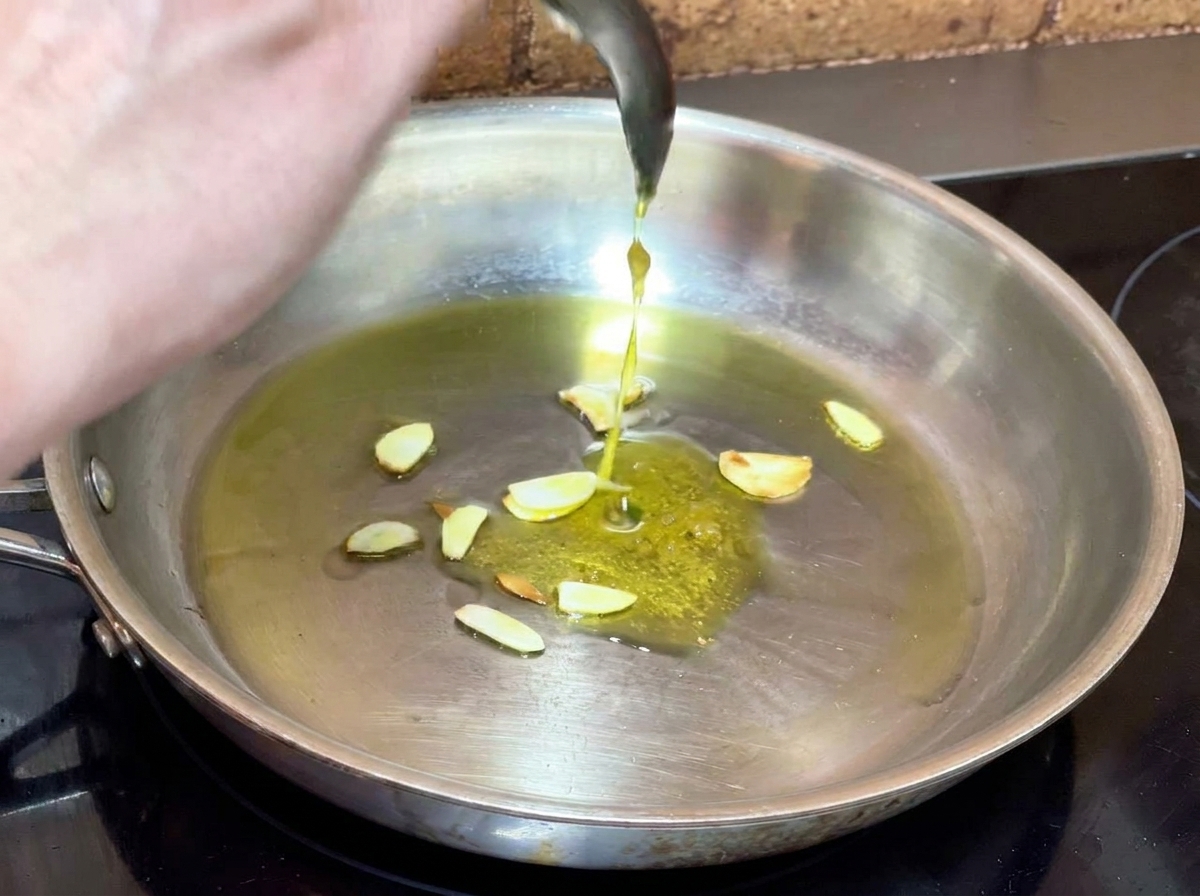 Cucchiaio che aggiunge la crema daglio preparata prima nella padella con lolio e laglio a fette.