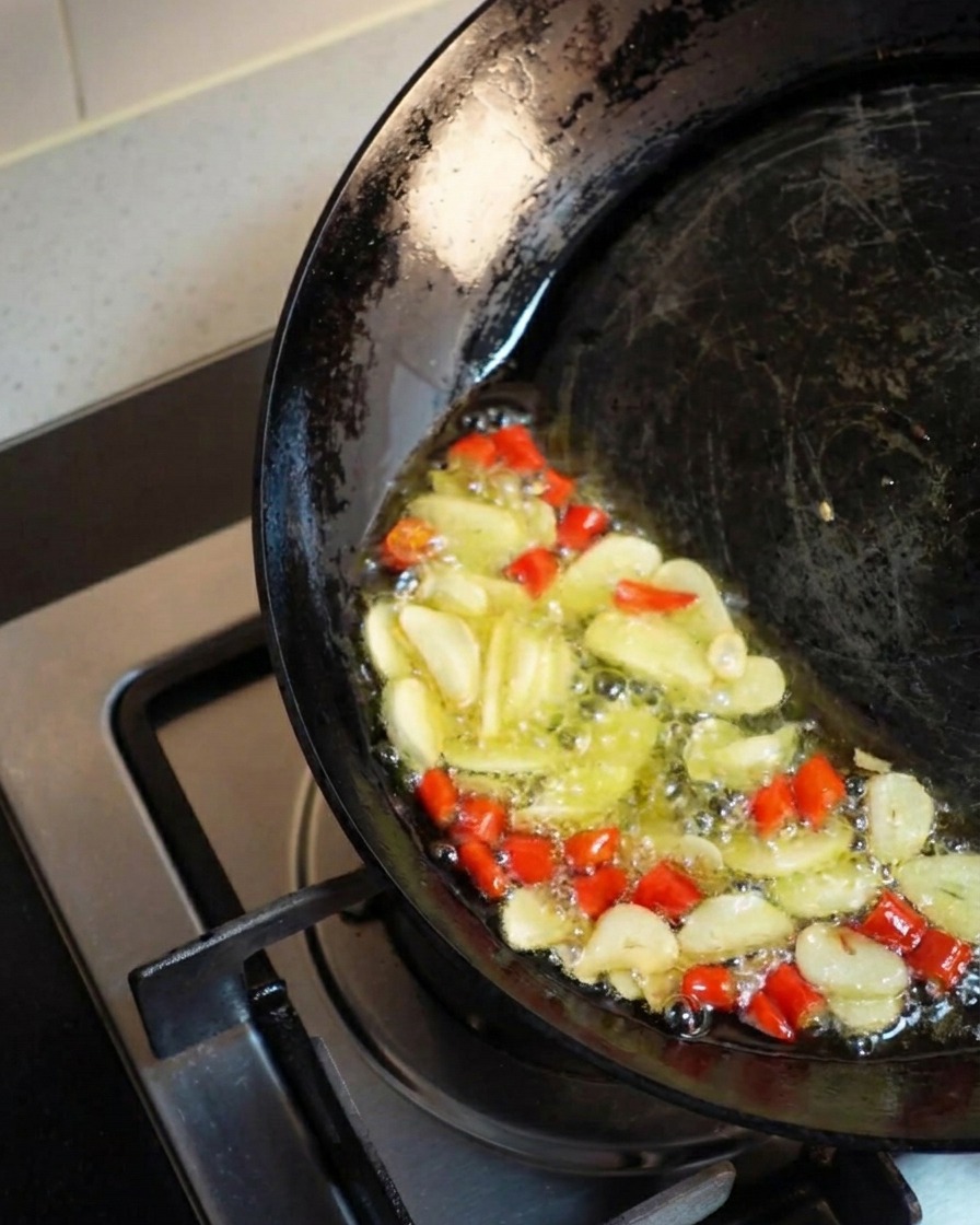 Un wok inclinato su un lato per raccogliere lolio dorato in cui aglio e peperoncino friggono vivacemente.