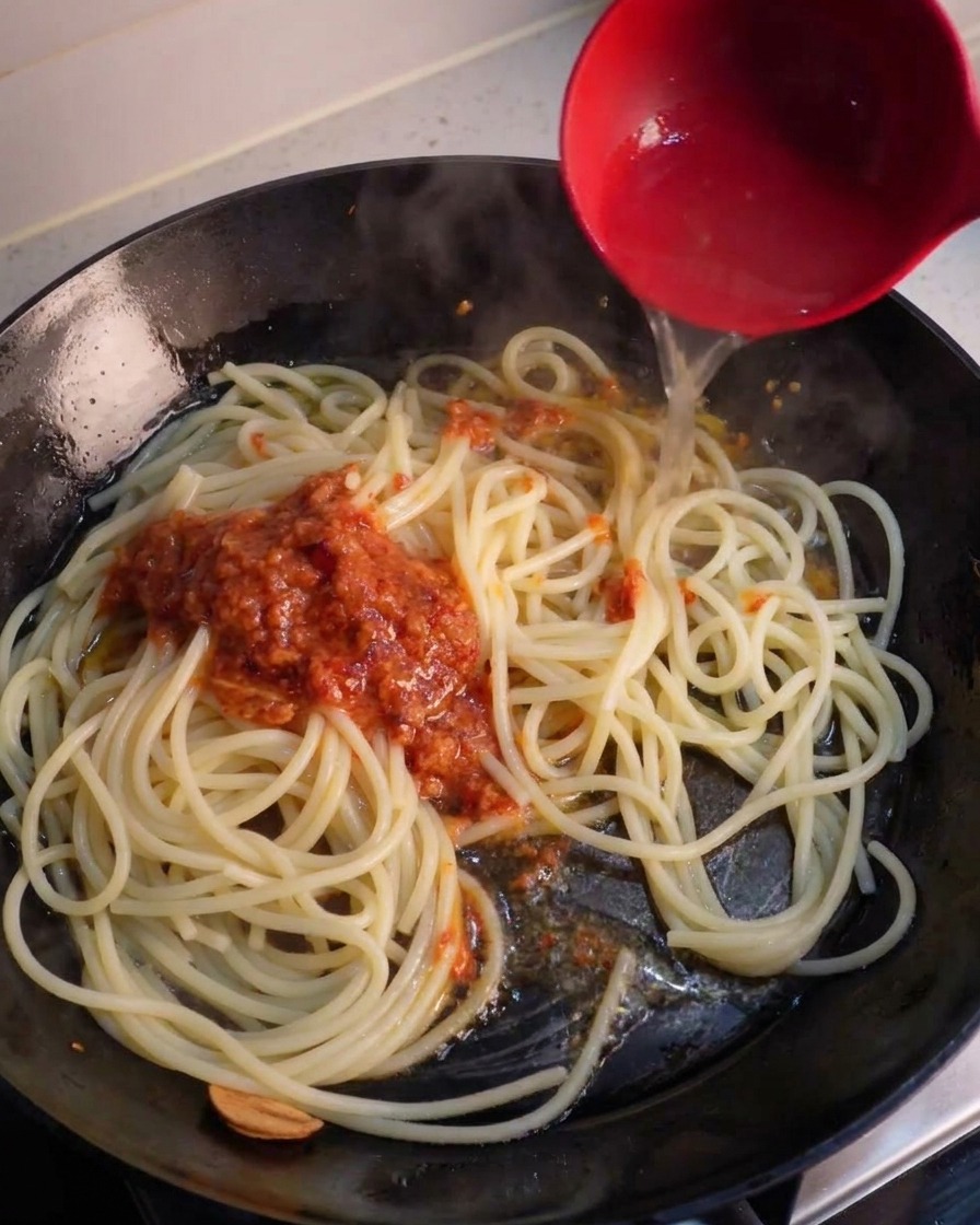 Un mestolo che versa acqua di cottura della pasta in padella sopra gli spaghetti e un cucchiaio di crema frullata al peperoncino.