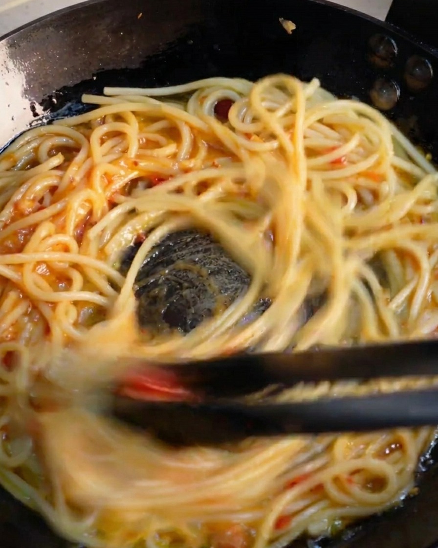 Pinze da cucina scure che saltano rapidamente gli spaghetti in padella per emulsionare la vibrante salsa allaglio e peperoncino.