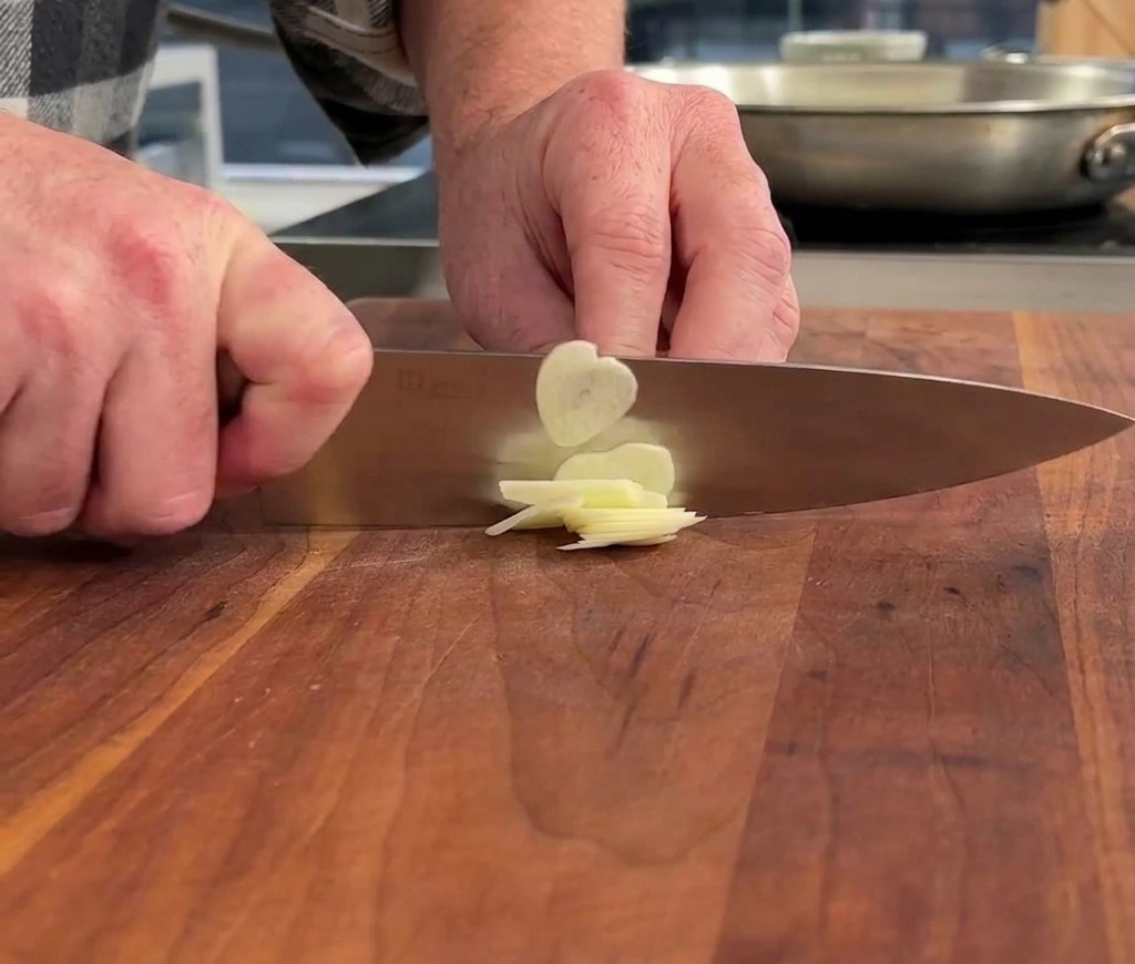Mani che usano un coltello da chef per affettare finemente uno spicchio daglio su un tagliere di legno.