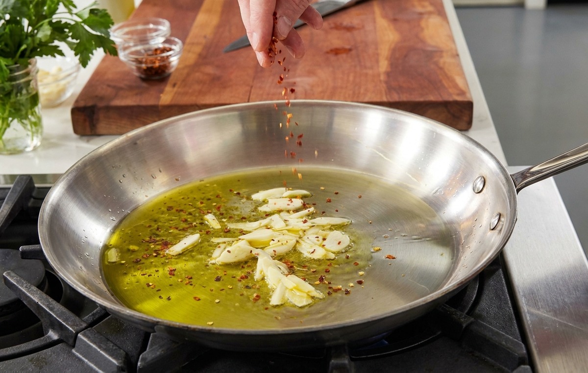 Una mano che cosparge scaglie di peperoncino in una padella con olio doliva e aglio a fette.