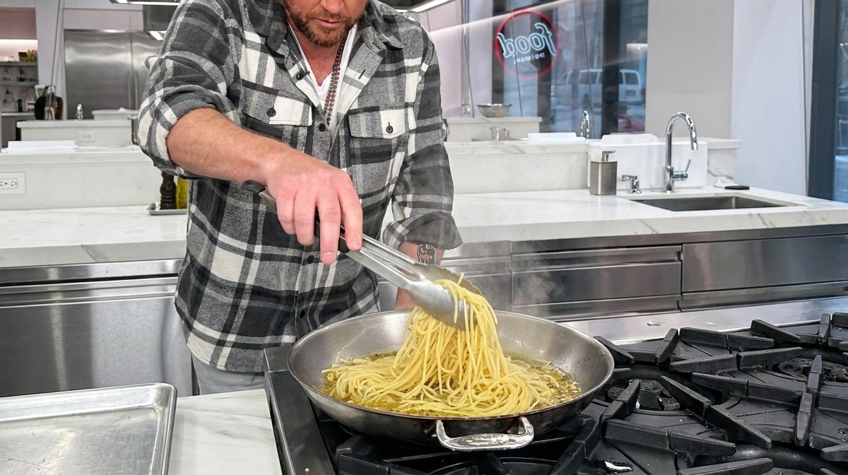 Spaghetti al dente che vengono tuffati in una padella calda con aglio, olio e acqua di cottura usando le pinze.