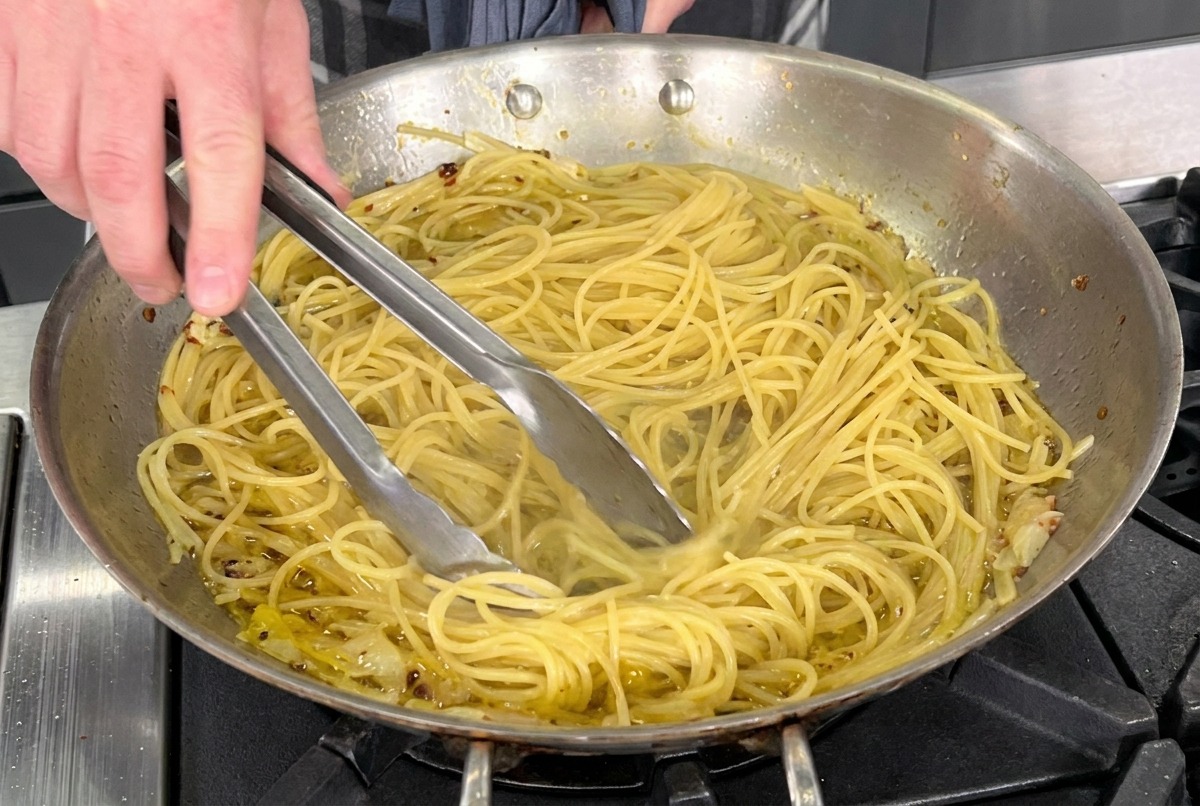 Pinze da cucina che saltano gli spaghetti in una padella di acciaio con olio, aglio e peperoncino.