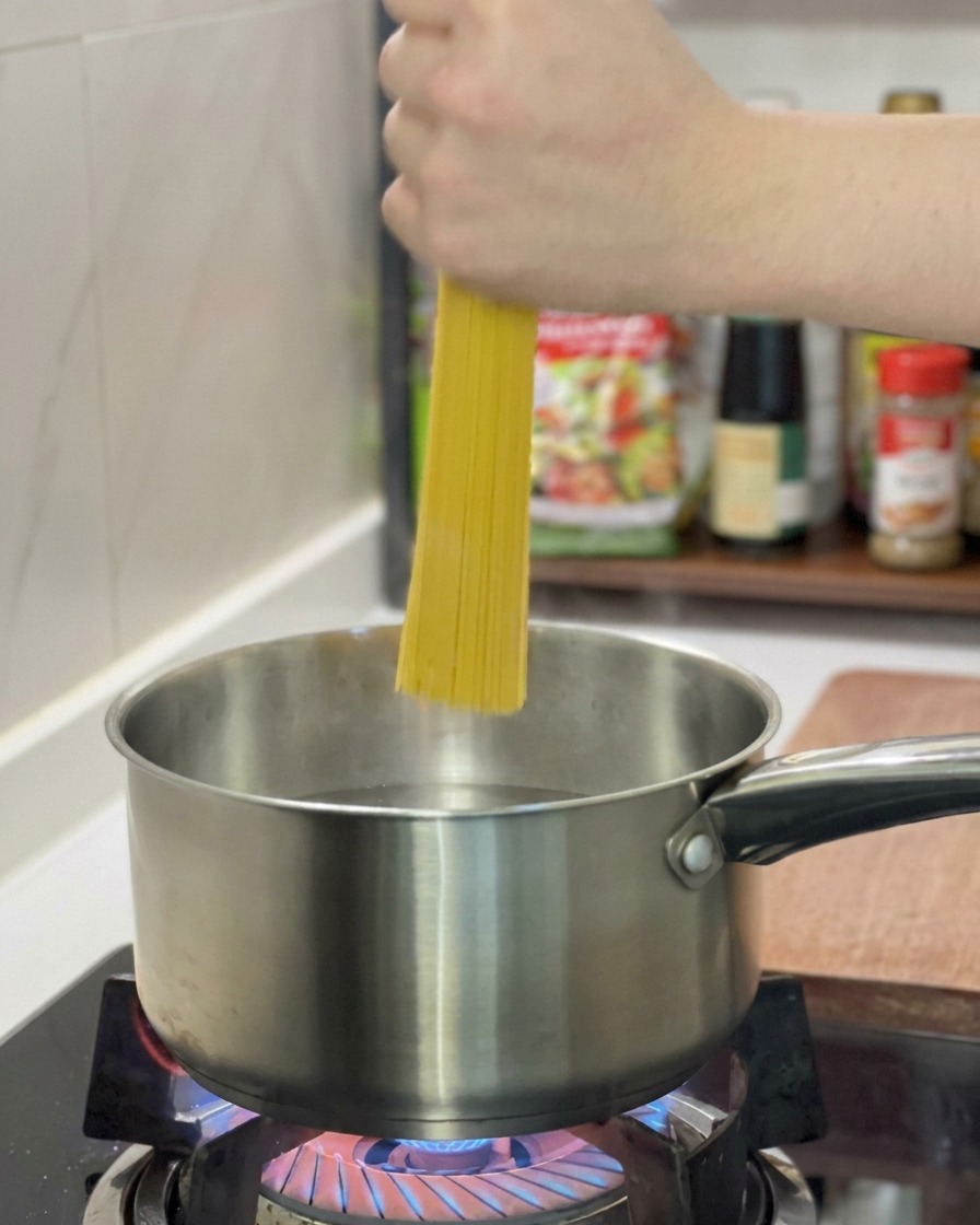 Una mano che tiene una manciata di spaghetti secchi in verticale mentre vengono immersi in una pentola dacciaio con acqua bollente.